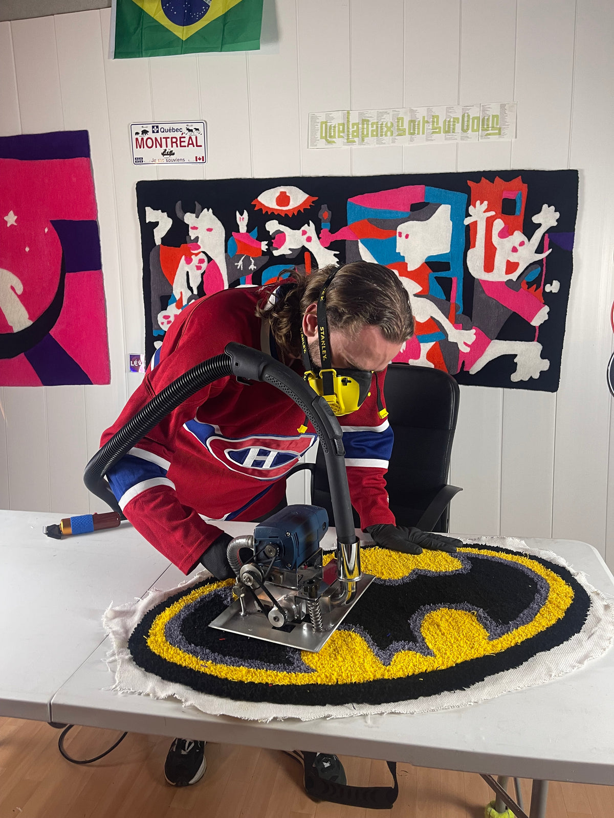 L'artisan de Signé Léon équipé d'un masque protecteur, réalisant les finitions minutieuses à la tondeuse professionnelle sur un tapis sur mesure représentant le logo Batman noir et jaune dans son atelier.