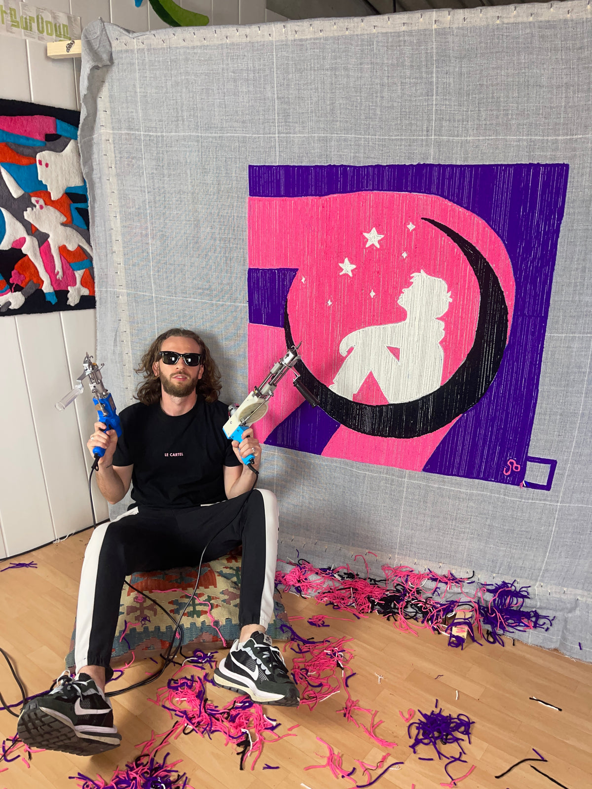 L'artisan de Signé Léon, souriant avec deux tufting guns en main, pose fièrement devant une grande et complexe création originale tuftée au motif géométrique coloré (rose, violet, noir). Son atelier est visible avec d'autres œuvres au mur et de la laine au sol.