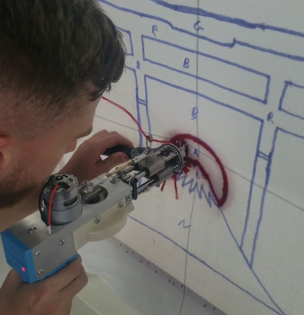 Plan rapproché d'un artisan concentré utilisant un pistolet de tufting mécanique pour insérer de la laine rouge épaisse sur une toile de base tendue. Il suit un motif géométrique complexe tracé en bleu avec des codes couleur visibles (lettres R, B, G, N) pour la fabrication d'un tapis sur mesure.