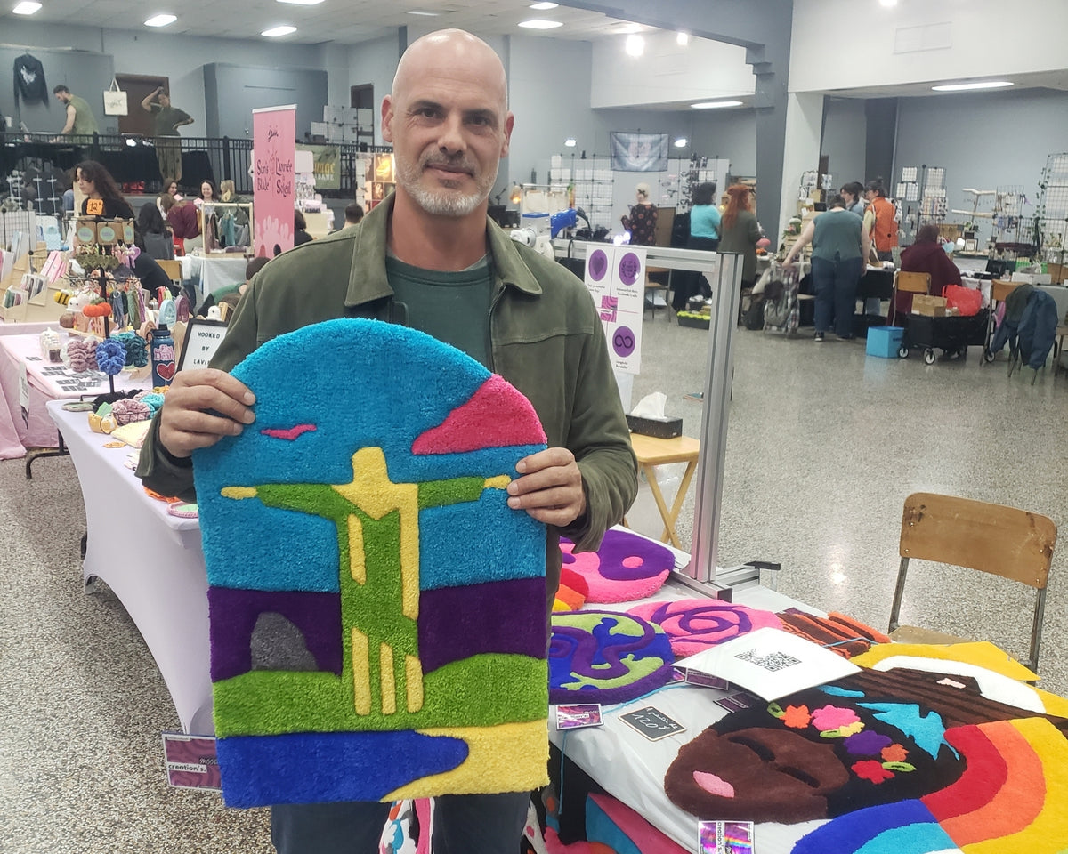 Un client ravi posant avec sa nouvelle acquisition sur le stand Signé Léon lors d'un salon de créateurs : une création originale tuftée à la main, très colorée, représentant le Christ Rédempteur.