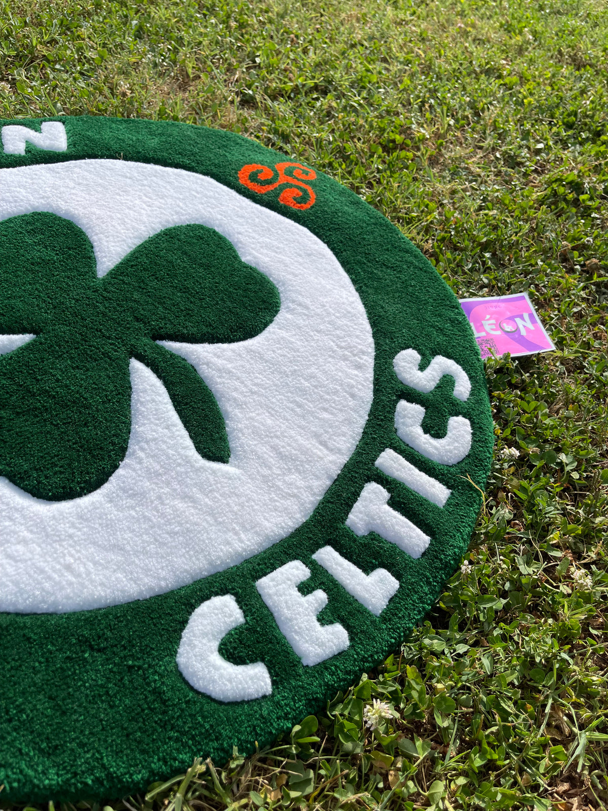 Détail de la typographie Celtics, des motifs celtiques orange et de l'étiquette artisanale Signé Léon sur le tapis tufté sur mesure.