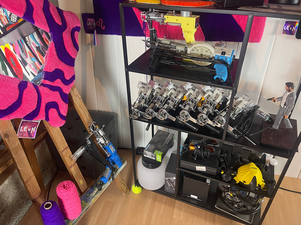 Rangement rempli de multiples tufting guns professionnels, de masques de protection et de matériel textile dans l'atelier de l'artisan Signé Léon. Sur la gauche, un grand miroir décoratif avec un cadre tufté rose et violet.