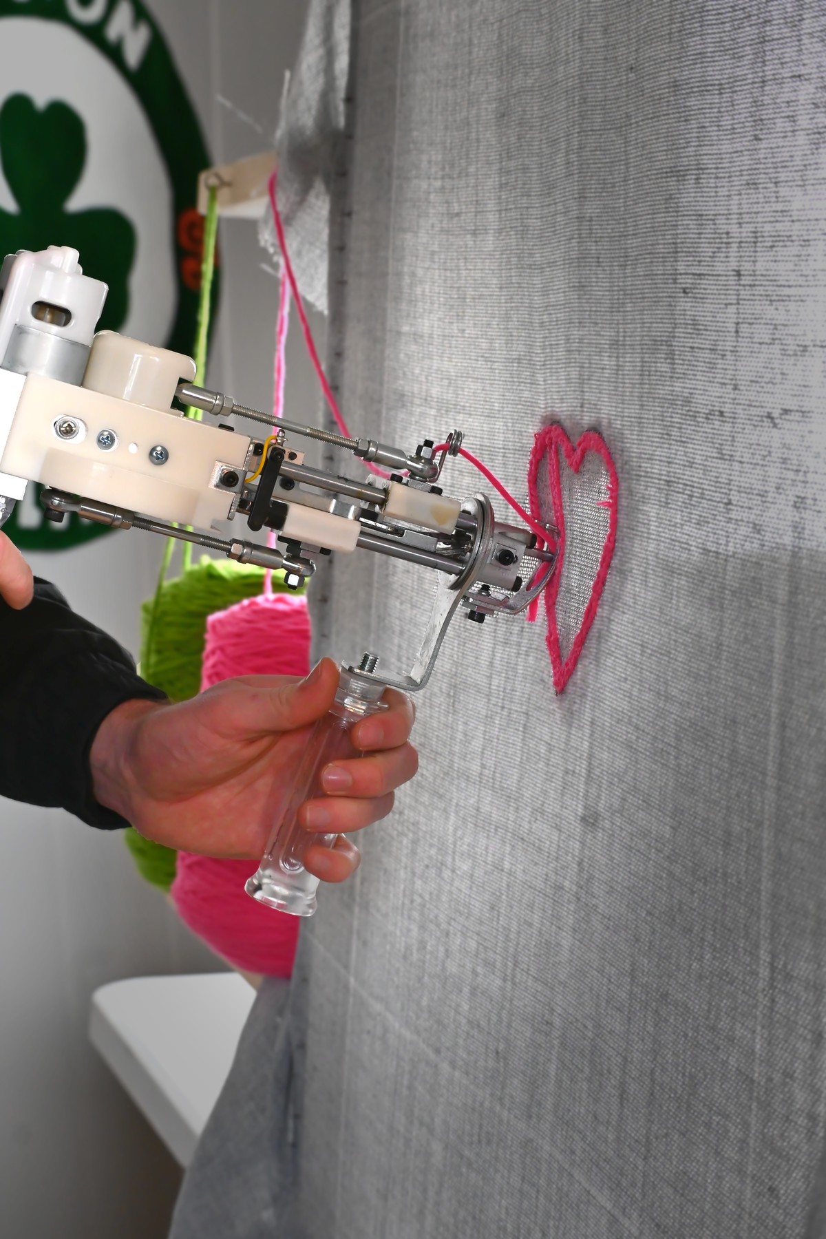 Gros plan détaillé sur la main de l'artisan Signé Léon maniant un pistolet à tufter pour insérer du fil de laine rose fluo et créer les contours d'un motif de cœur sur une toile de fond grise.