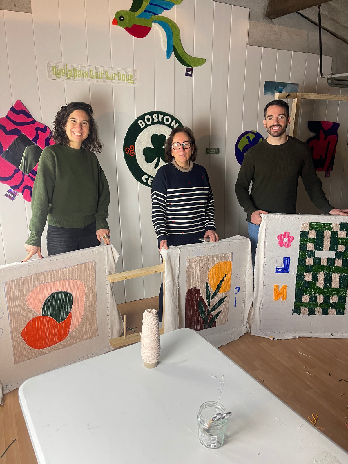 Trois participants souriants présentant fièrement leurs tapis tuftés sur des cadres à la fin d'une initiation dans l'atelier Signé Léon, près de Toulouse. En arrière-plan, le mur de l'atelier est décoré de plusieurs créations textiles originales.