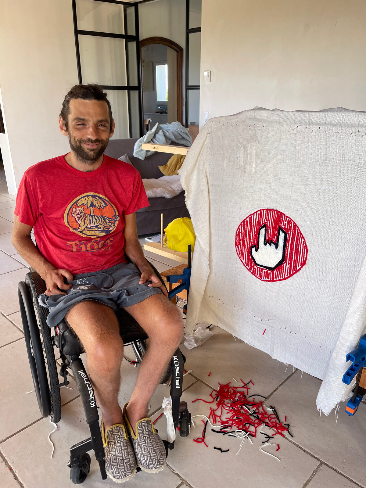 Homme souriant en fauteuil roulant devant son tapis en cours de création lors d'une initiation au tufting à domicile encadré par l'artiste Tufting Signé Léon.