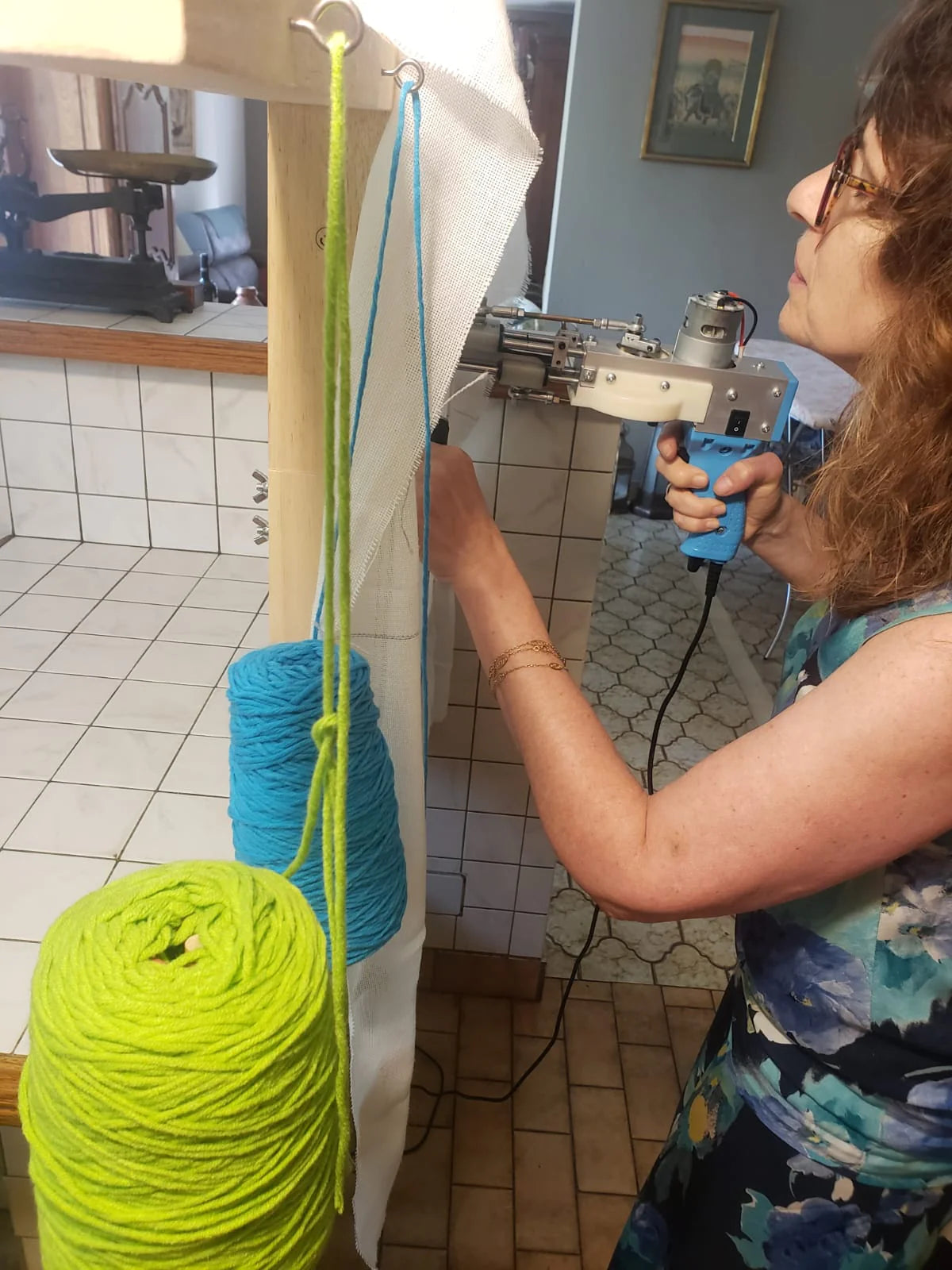 Une participante concentrée maniant le tufting gun sur sa toile tendue, avec des bobines de laine verte et bleue, lors d'un atelier de création de tapis à domicile.