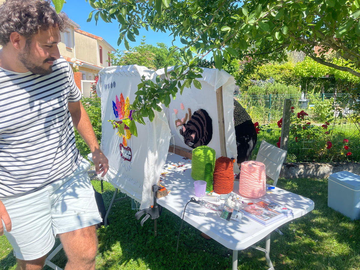 Un participant observant sa création lors d'une initiation au tufting en plein air, dans le jardin ensoleillé de l'atelier Signé Léon près de Toulouse. Des cadres et des pelotes de laine colorées sont installés sur une table sous les arbres.