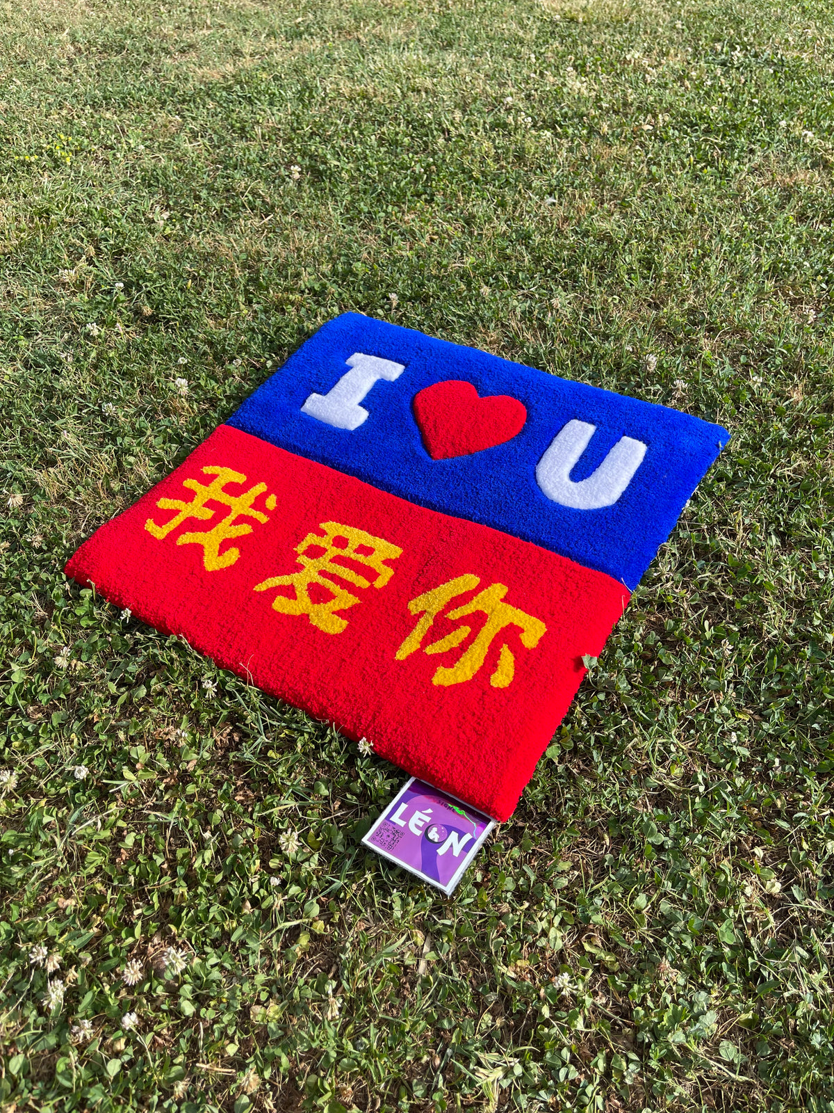 Vue de biais du tapis tufté Amour sur la pelouse, mettant en valeur l'harmonie visuelle entre les deux langues emblématiques et le volume de la laine.