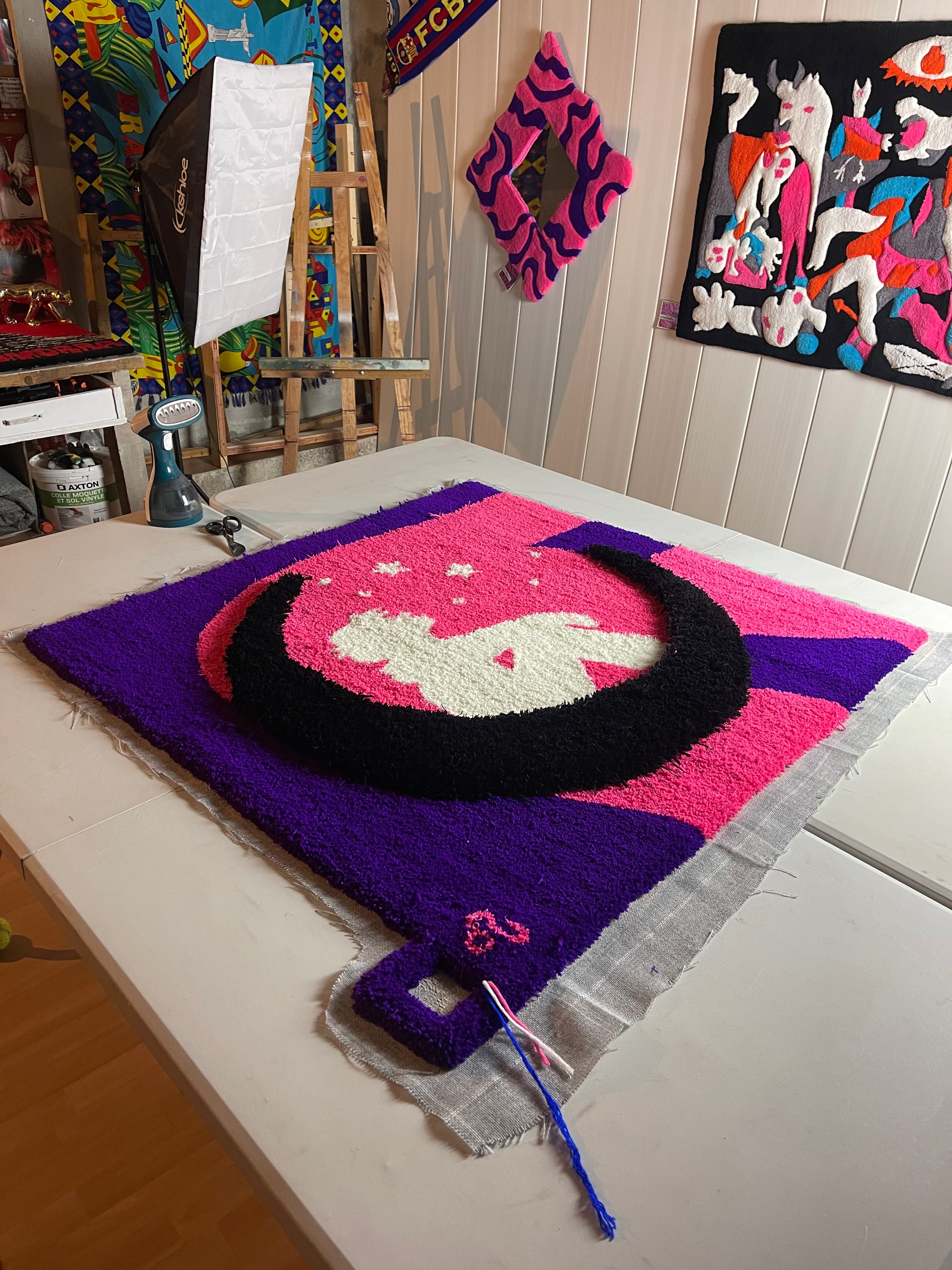 Tapis sur mesure en cours de finition sur la table de l'atelier Signé Léon, représentant une lune noire et une silhouette sur un fond rose et violet vibrant.