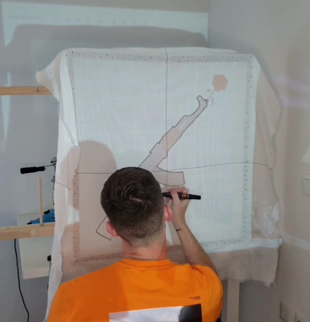 L'artisan Signé Léon vu de dos, en train de décalquer minutieusement au marqueur noir un motif projeté (un fusil avec une rose) sur une toile tendue. Première étape préparatoire pour la création d'un tapis tufté sur mesure.