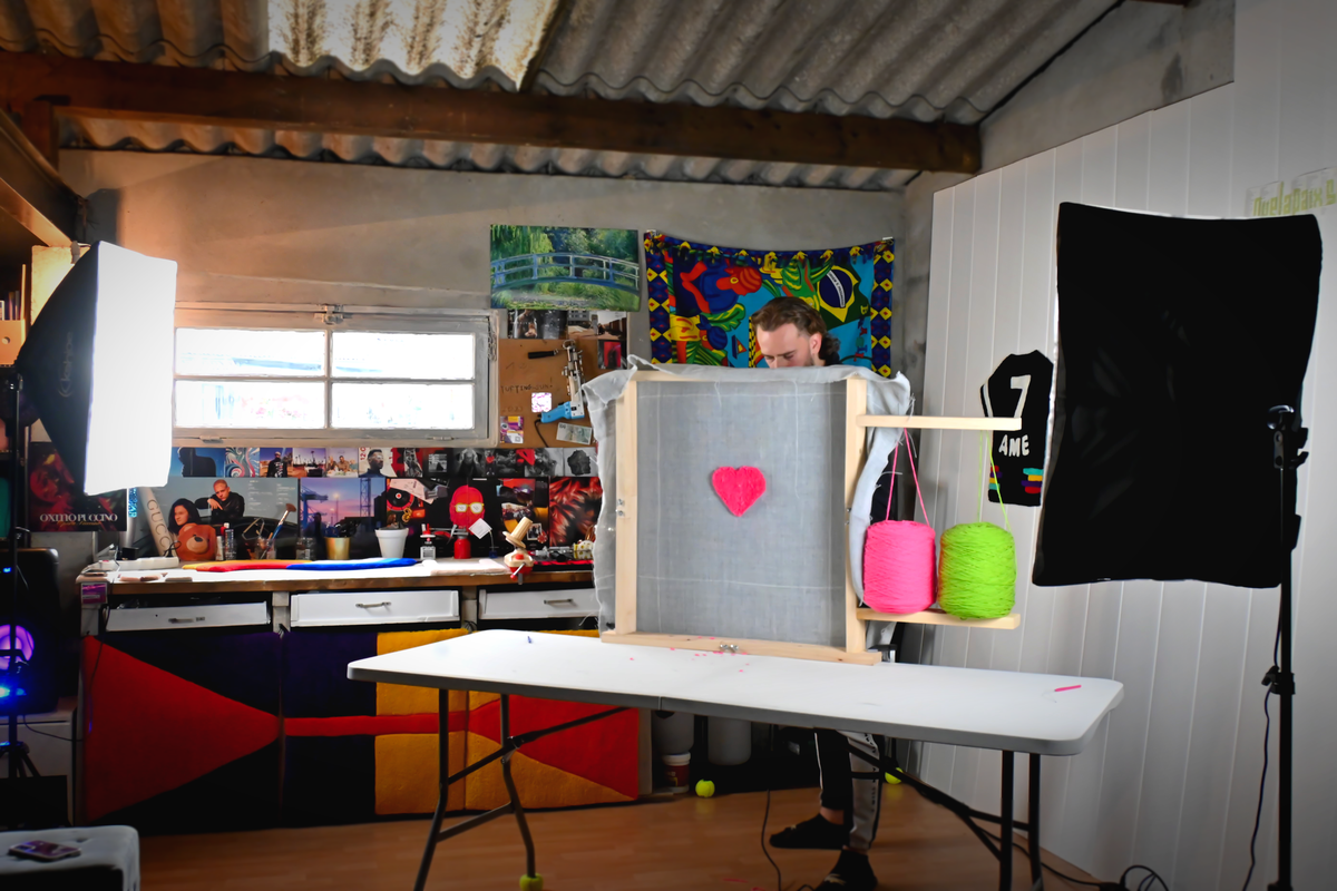 L'artisan de Signé Léon travaillant sur un cadre de tufting dans son atelier, en train de créer un motif de cœur rose. L'espace est aménagé avec des projecteurs photo professionnels et décoré de nombreuses inspirations murales.